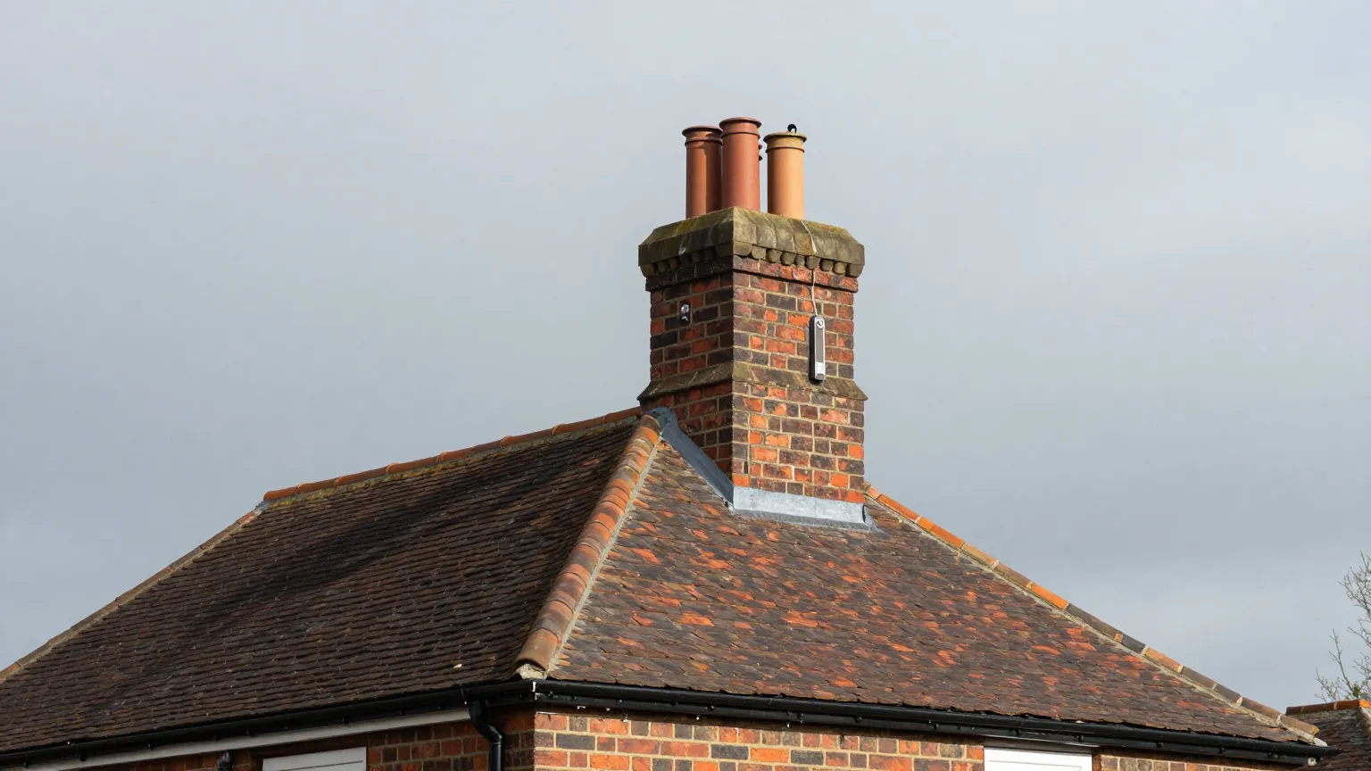 A tiled hip roof with a red brick chimney stack and three clay chimney pots against a grey sky